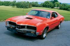 an orange muscle car parked in a parking lot next to some grass and trees on a sunny day