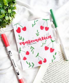 two pens and an open book on top of a bed next to some flowers, leaves and a plant