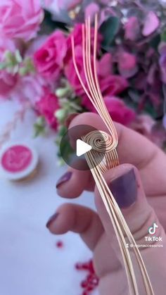 a woman is holding a piece of metal wire in her hand with pink flowers behind it