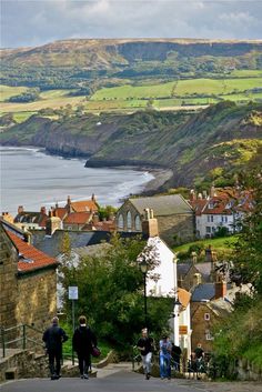 some people are walking down a hill by the water and buildings with hills in the background