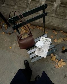 a person sitting on a bench reading a newspaper and holding a brown handbag next to them