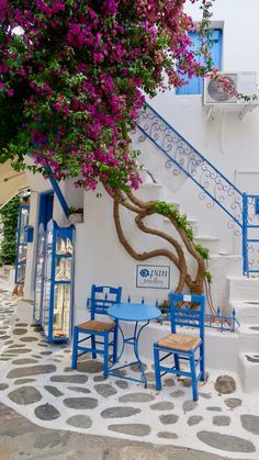 a blue table and chairs under a tree with purple flowers in front of a white building