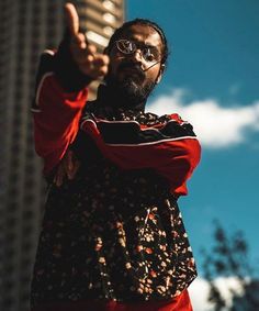 a man with glasses and a red jacket pointing at something in the air while standing next to a tall building