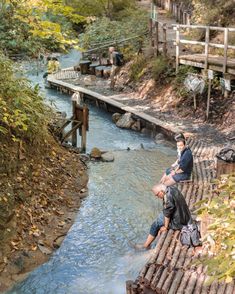 two men sitting on the edge of a wooden bridge over a river with blue water