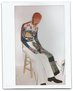 a man sitting on top of a wooden chair next to a white wall wearing a red helmet