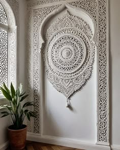 an ornate white wall hanging next to a potted plant in front of a window