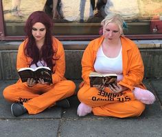 two women are sitting on the ground reading books while wearing orange jumpsuits and holding teddy bears