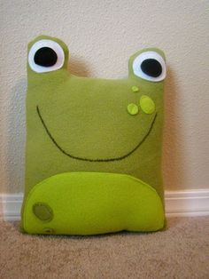 a green frog pillow with eyes and nose sitting on the floor next to a wall