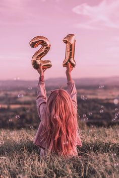 a girl with pink hair is holding up two gold balloons in the shape of numbers