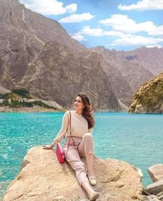 a woman sitting on top of a large rock next to a blue body of water