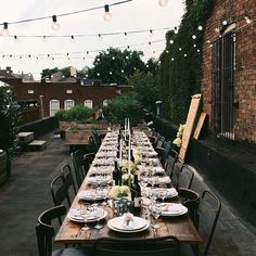 an outdoor dining table set up with place settings and lights strung from the ceiling above