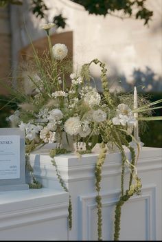 white flowers and greenery are in a vase on a mantle with an address card