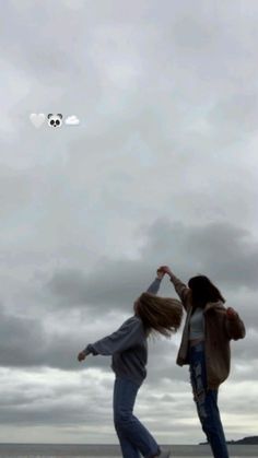 two young women are flying a kite on the beach in front of an overcast sky