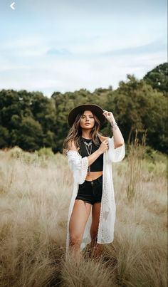 a woman standing in tall grass wearing a black hat and white cardigan sweater over her shoulders