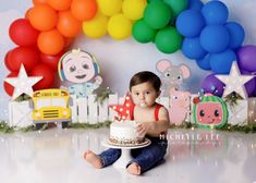 a baby sitting on the floor with a cake and balloons in front of him,