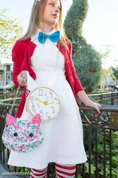 a woman in a white dress and red cardigan holding a clock with her face painted like a cat