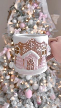 someone holding up a coffee mug with a gingerbread house on it in front of a christmas tree