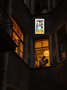 two people standing in an open window looking at the light from below them on a building