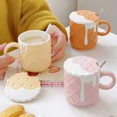 a person holding a cup and some cookies on a table with two mugs in front of them