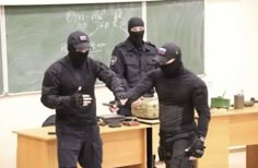 three men in black jackets and masks standing near a desk