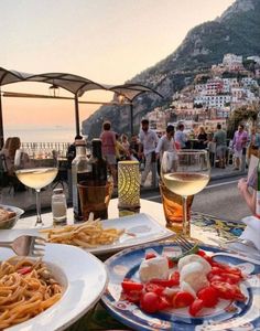 plates of food and glasses of wine on a table overlooking the ocean with people standing around