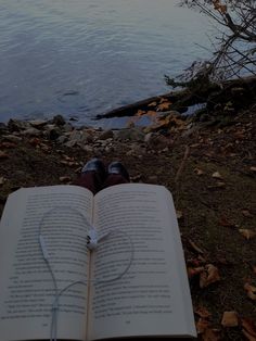 an open book sitting on the ground next to a body of water with trees and leaves around it