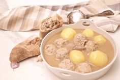 a bowl filled with meat and potatoes next to bread on a white tableclothed cloth