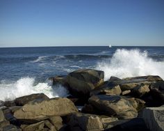 the waves are crashing on the rocks by the ocean's shore as a sailboat is in the distance