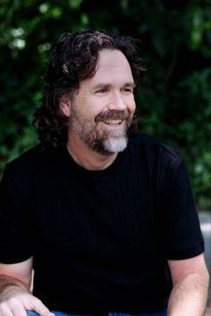 a man with long hair and beard sitting on a bench in front of some trees