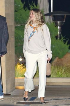 a woman in white pants and a gray sweater is walking down the street at night