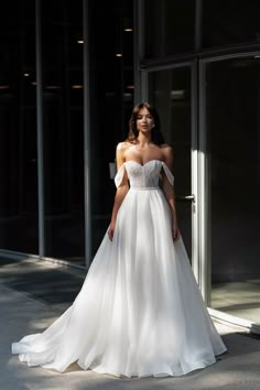 a woman standing in front of a building wearing a wedding dress with an off the shoulder neckline