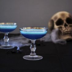 two glasses with blue liquid in front of a skull