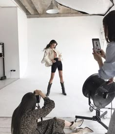 a woman in black shorts and boots is taking a photo with her cell phone while two other people are sitting on the floor