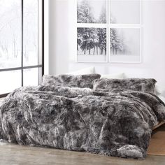 the bed is made with grey fur and pillows on top of wooden flooring in front of large windows