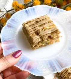 a hand holding a white plate with food on it and flowers in the back ground