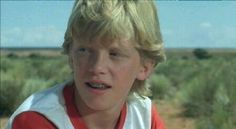 a young boy with blonde hair wearing a red and white shirt in front of a desert landscape