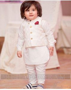 a little boy wearing a white suit and bow tie standing in front of a table