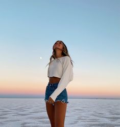 a woman standing on top of a sandy beach next to the ocean at sunset with her eyes closed