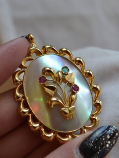 This colourful, whimsical and playful vintage brooch is made with a flowing, scalloped, ribbon-like border of 14kt yellow gold-plated rose gold; it swirls effortlessly around a beautifully hued, rainbow-life, super lustrous mother of pearl inset, which has been further set with an adorable, sculptural gold floral motif. In three chunky, lovely bezels protruding artfully from the surface, two synthetic rubies and a synthetic emerald glint like bright candies. It measures 4.5 CM in length, NOT inc