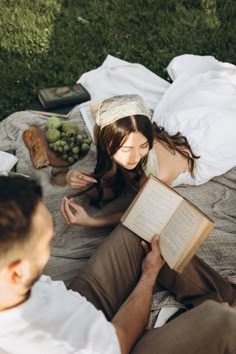 a woman reading a book while sitting on a blanket in the grass next to a man