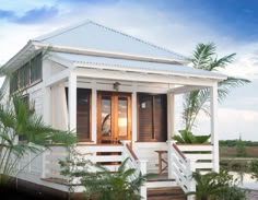 a small white house with brown shutters and wooden doors on the front porch is surrounded by palm trees