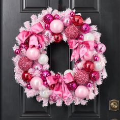 a pink and white christmas wreath on a black door with ornaments hanging from the front