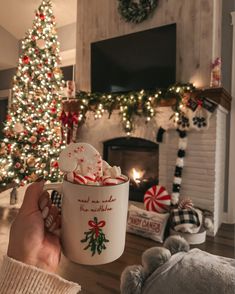 a person holding a coffee mug in front of a fireplace with christmas decorations on it