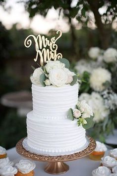 a wedding cake with white flowers and mr and mrs topper on the table next to cupcakes