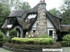 a small house with a stone chimney in the front yard and bushes around it, surrounded by greenery