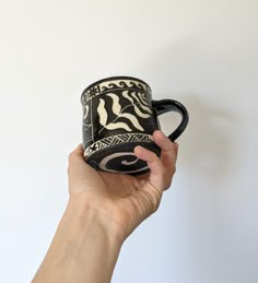 a hand holding a black and white coffee mug with designs on the outside, in front of a white wall