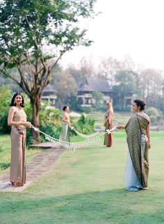 two women in sari are holding hands with each other on the grass near some trees