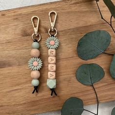 two keychains with flowers and letters on them sitting on a wooden table next to leaves