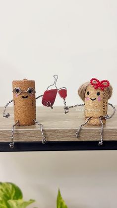 two corks with faces and arms are holding wine glasses on a wooden shelf next to a potted plant