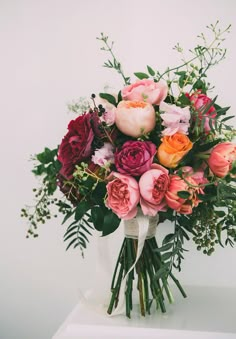 a bouquet of colorful flowers in a white vase on top of a table with greenery